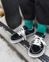 Person wearing black SPLAY REV LTS sneakers and green socks beside a skateboard.