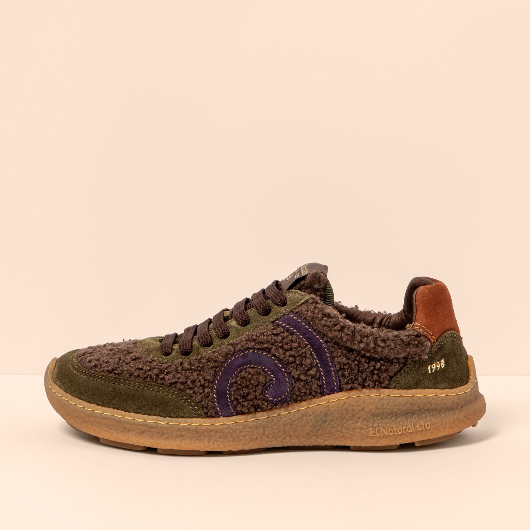 Zapatilla marrón de perfil, con tejido rizado, refuerzos verde oliva y espiral morada.